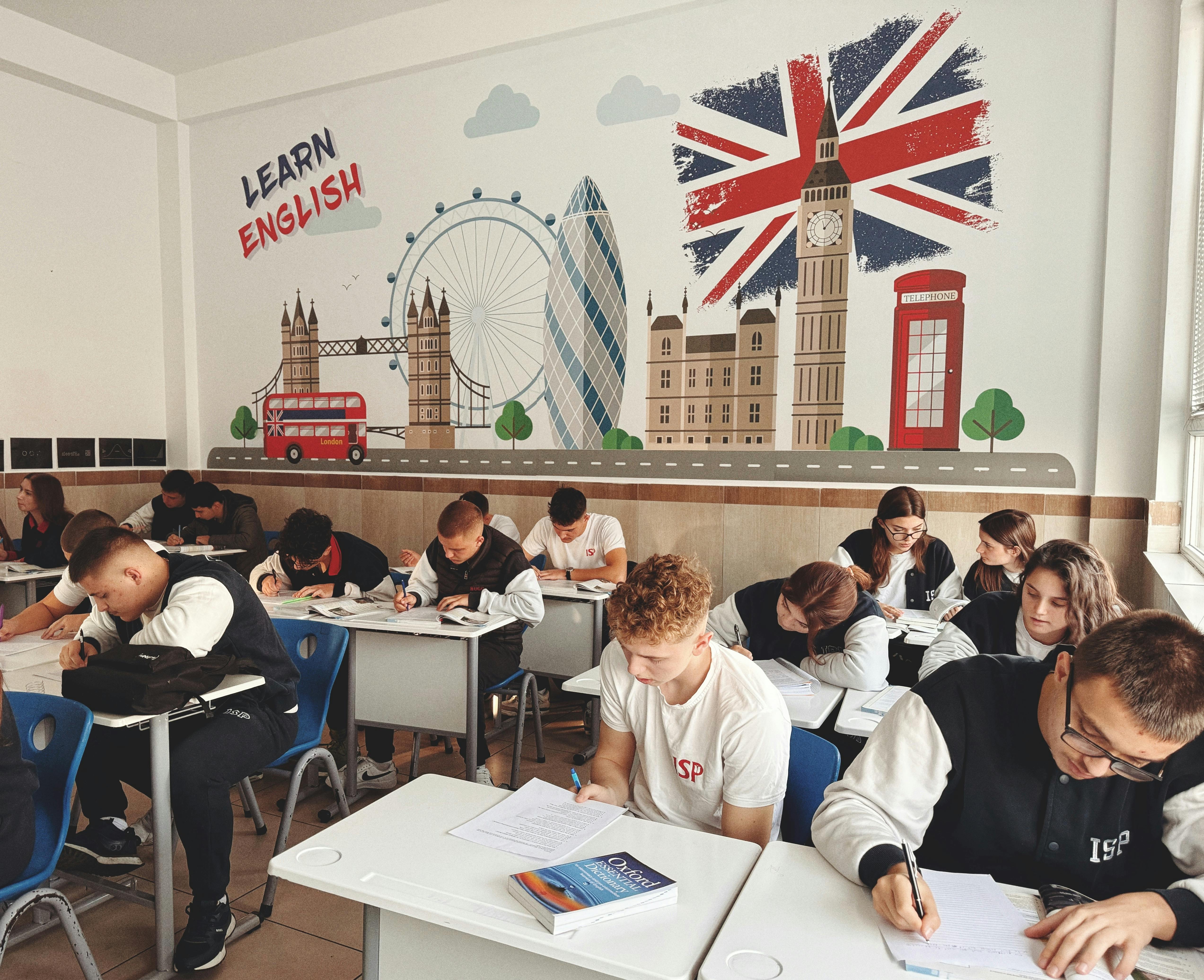 Cursos e inmersiones lingüísticas en inglés para el verano. Estudiantes en un aula con un mural de Londres, ilustrando los programas de inmersión lingüística en España y campamentos de inglés en el extranjero.
