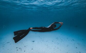 Una persona equipada con traje de neopreno y aletas suspendida horizontalmente bajo el agua, ilustrando las prácticas de dominio de la flotabilidad en submarinismo.