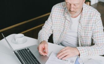 Un hombre jubilado revisando facturas y el borrador de la renta en su portátil, ilustrando si un jubilado debe hacer la Declaración de la Renta según sus ingresos.