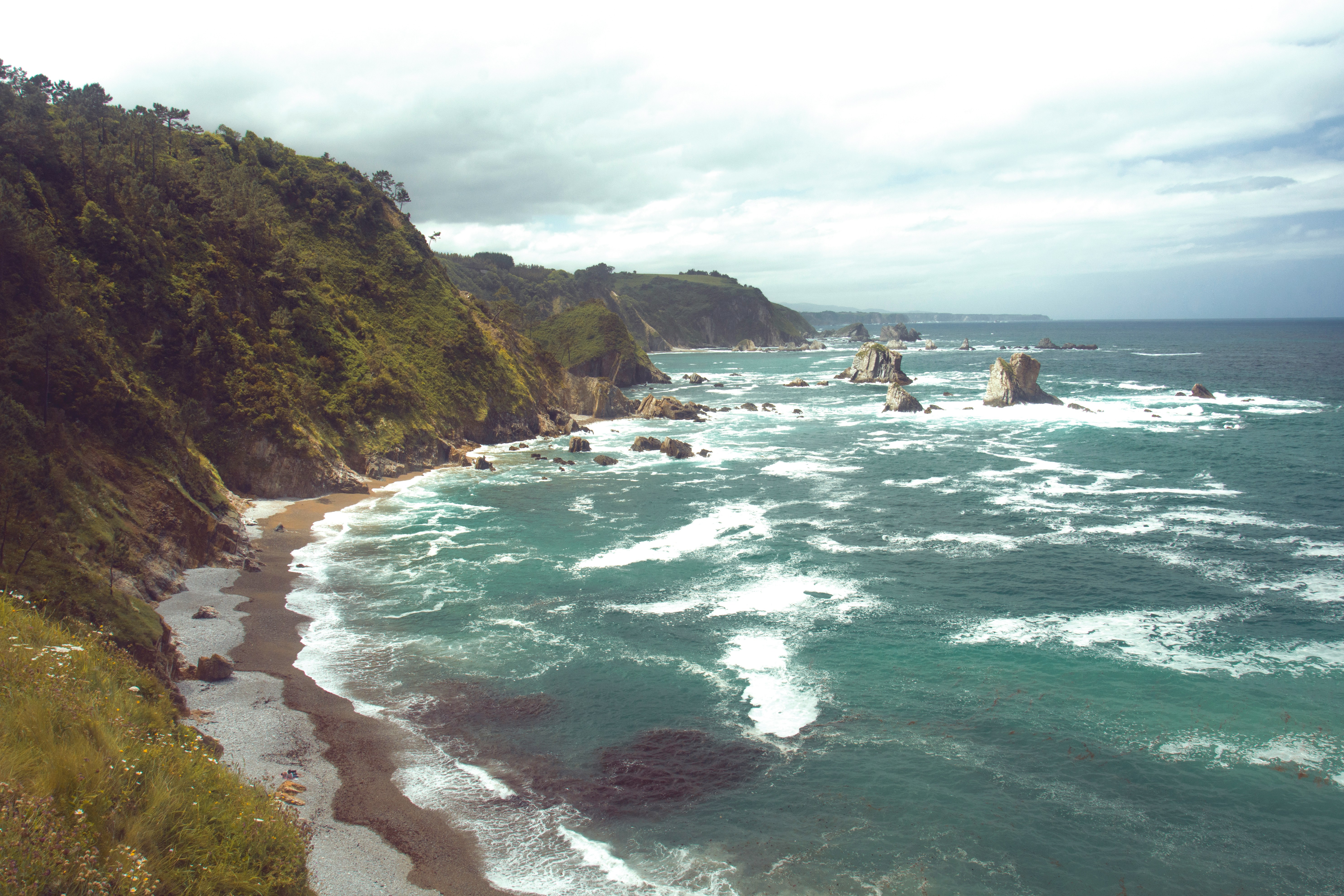 Entorno natural y acantilados de la Playa del Silencio. Vista panorámica desde la cima de un acantilado verde hacia la Playa del Silencio en Asturias, mostrando la costa rocosa, islotes y el mar Cantábrico con espuma blanca.