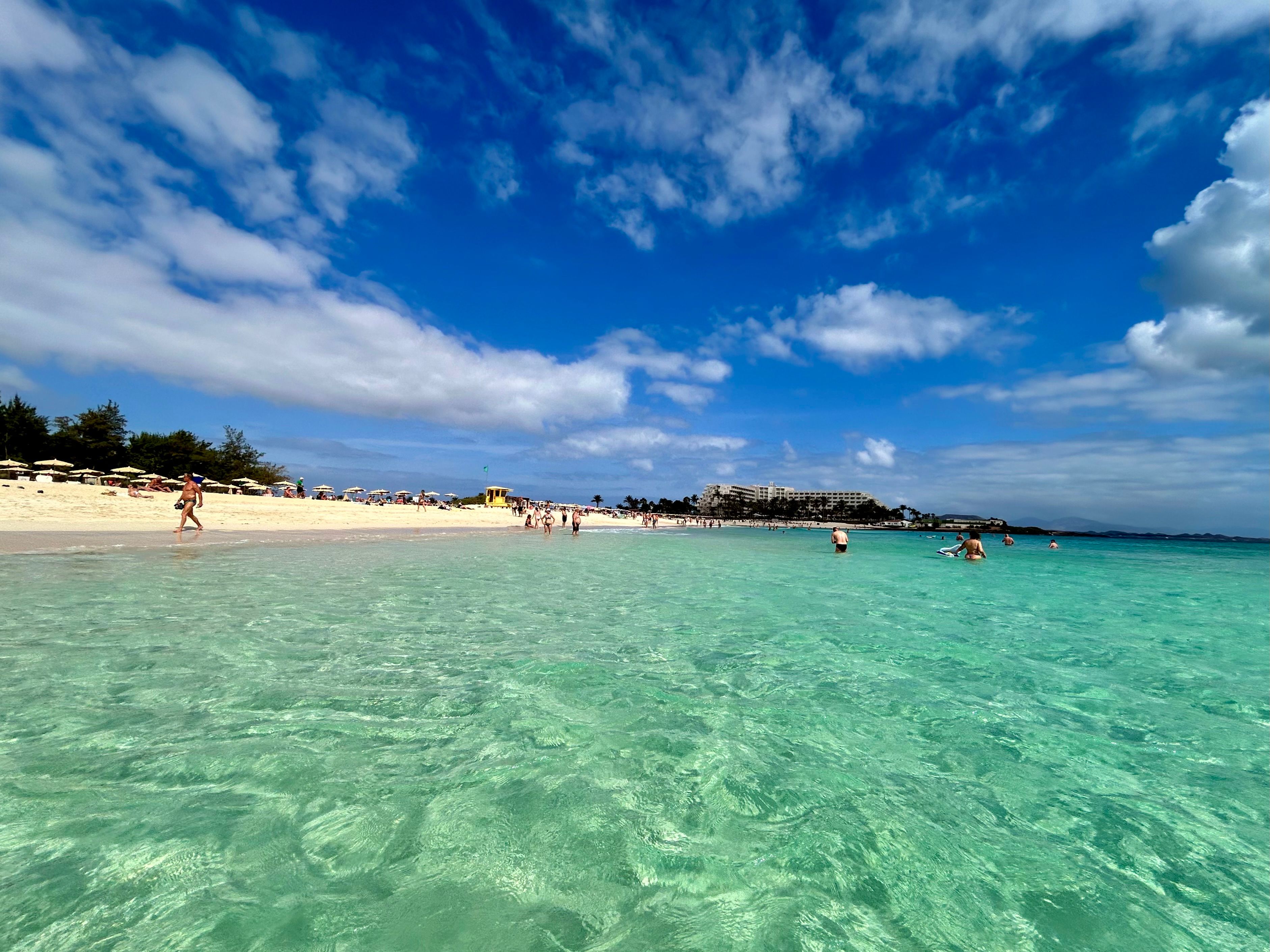 Aguas cristalinas y arena fina en las playas de Corralejo. Bañistas en la orilla de las Grandes Playas de Corralejo en Fuerteventura, con aguas turquesas y transparentes bajo un cielo azul con nubes.