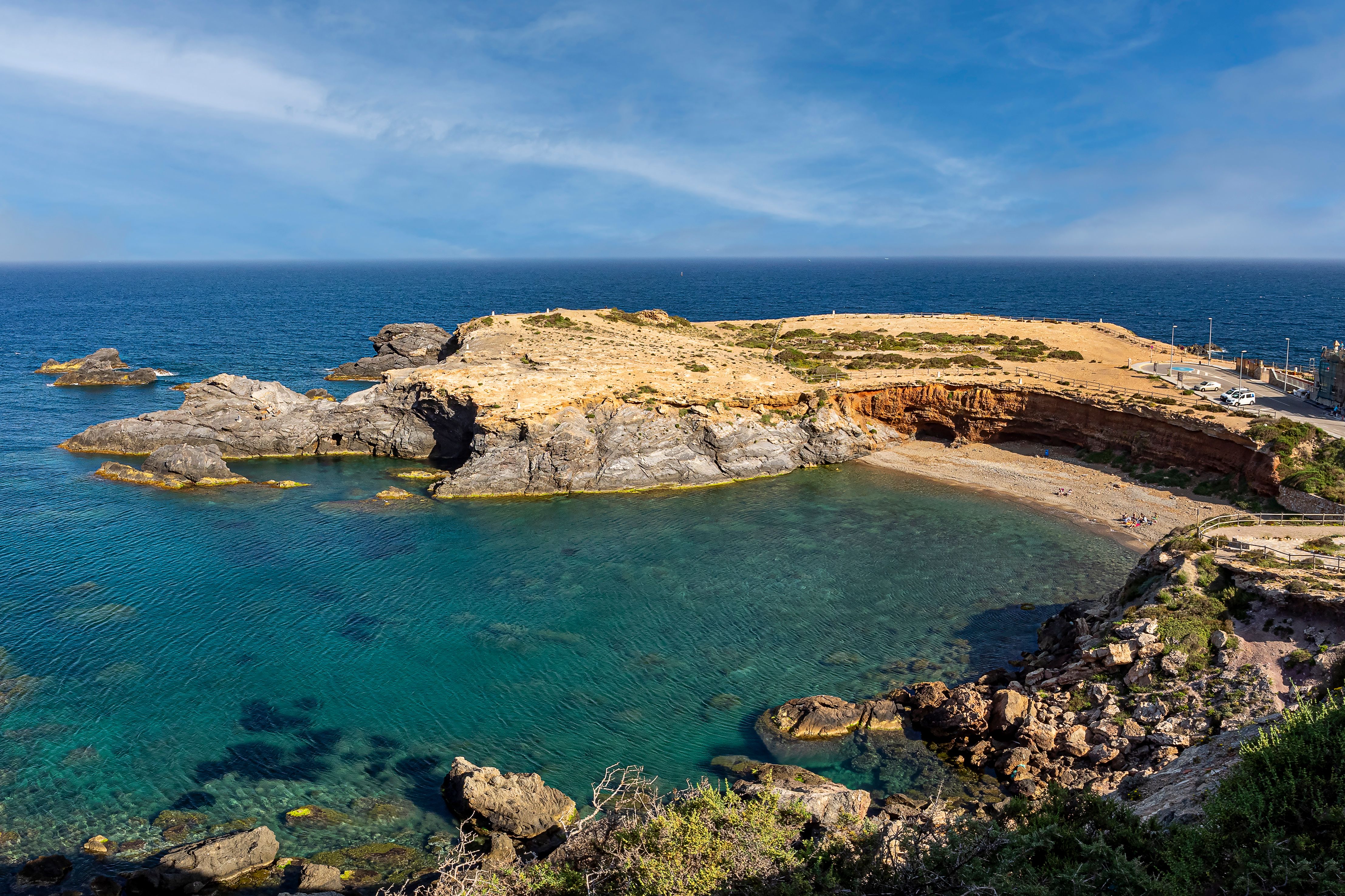 Paisaje costero y formaciones geológicas en la Playa de Calblanque. Vista panorámica elevada de una cala protegida por formaciones rocosas y acantilados con agua cristalina, ilustrando el entorno virgen del Parque Regional de Calblanque en Cartagena.