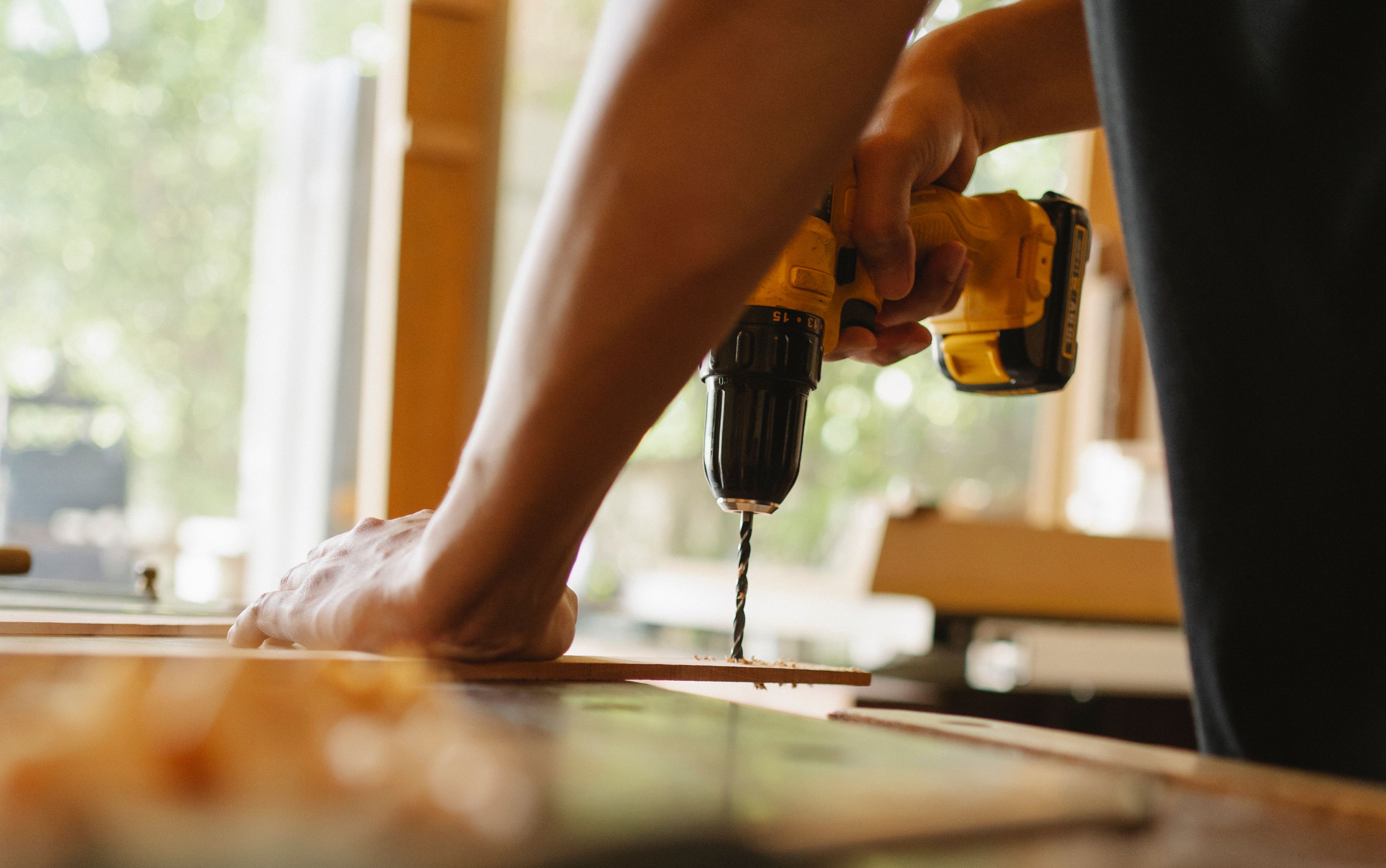 Prestaciones y coberturas de la baja por contingencia profesional o laboral. Primer plano de las manos de un operario utilizando un taladro eléctrico inalámbrico sobre una plancha de madera, ilustrando los requisitos para tramitar una baja por accidente de trabajo.