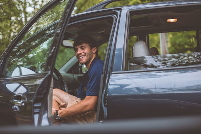 Chico joven sonriendo sentado dentro del coche con la puerta abierta